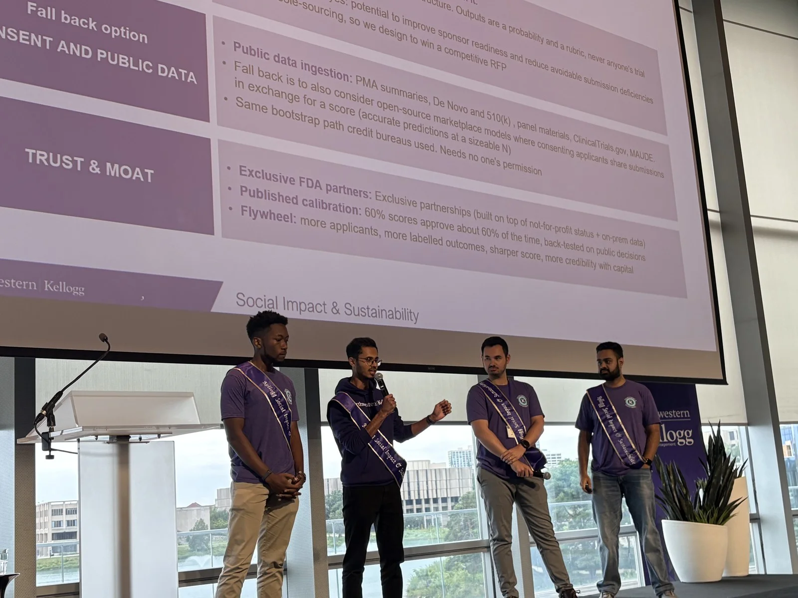 The four MedScore teammates presenting on stage at Kellogg, with their proposal projected behind them